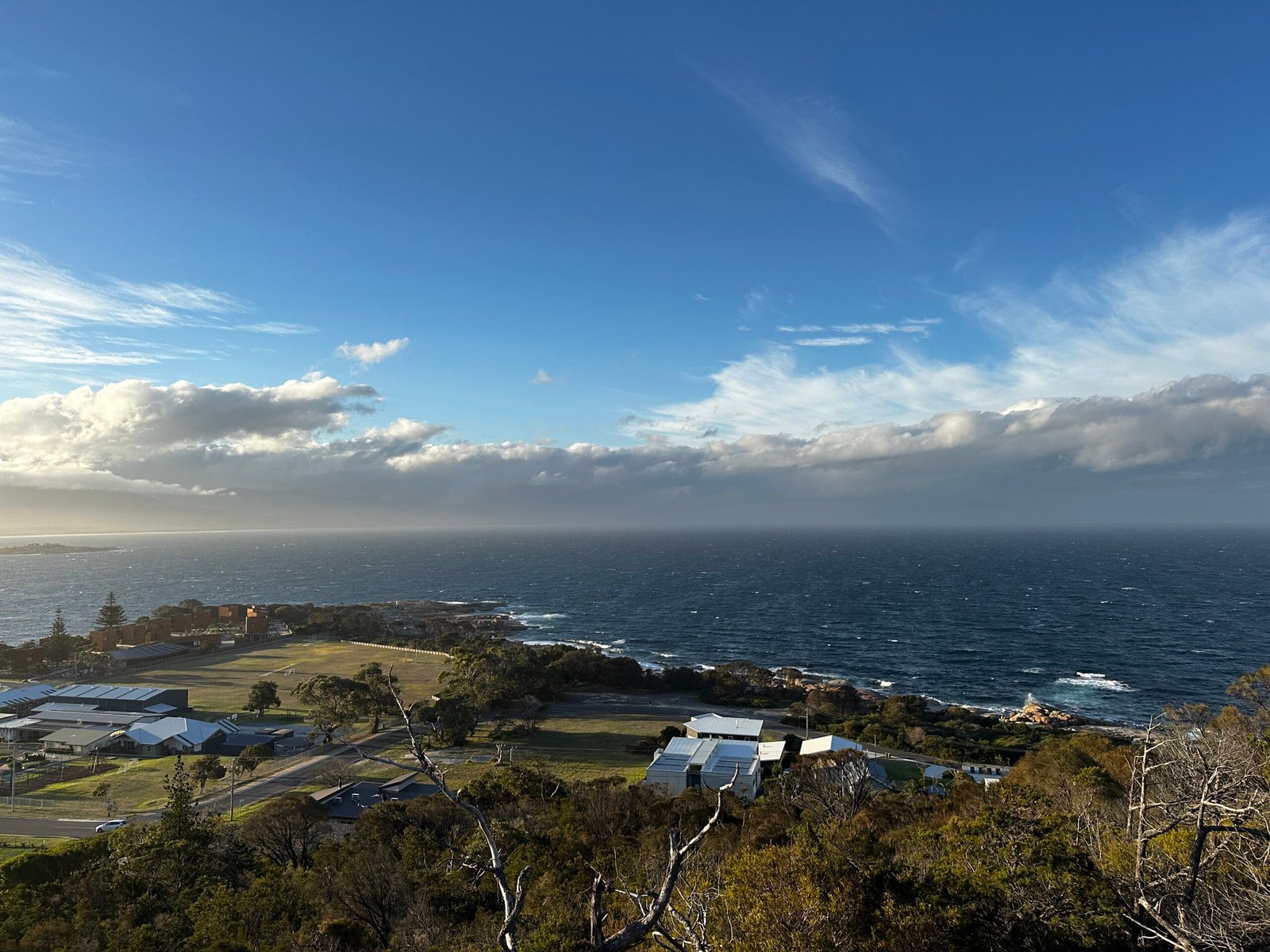 Coastal town seen from above, with low houses, open fields, and a deep blue ocean under a wide sky with scattered clouds.